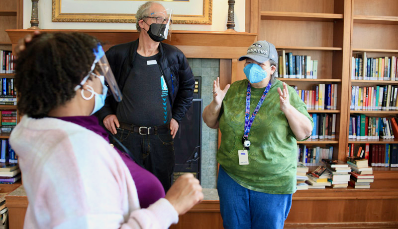A group laughing in a library in front of a fireplace. Everyone is wearing face masks, and the Ombuds are wearing face shields. The two in face shields are focused on a person in green shirt gesturing with a sparkle in their eye telling a joke..