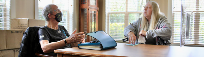 Two people speaking at a table one has long blonde hair and is sitting in a wheelchair the Ombuds has an open folder in front of him and is gesturing while wearing a t-shirt, face shield, and face mask