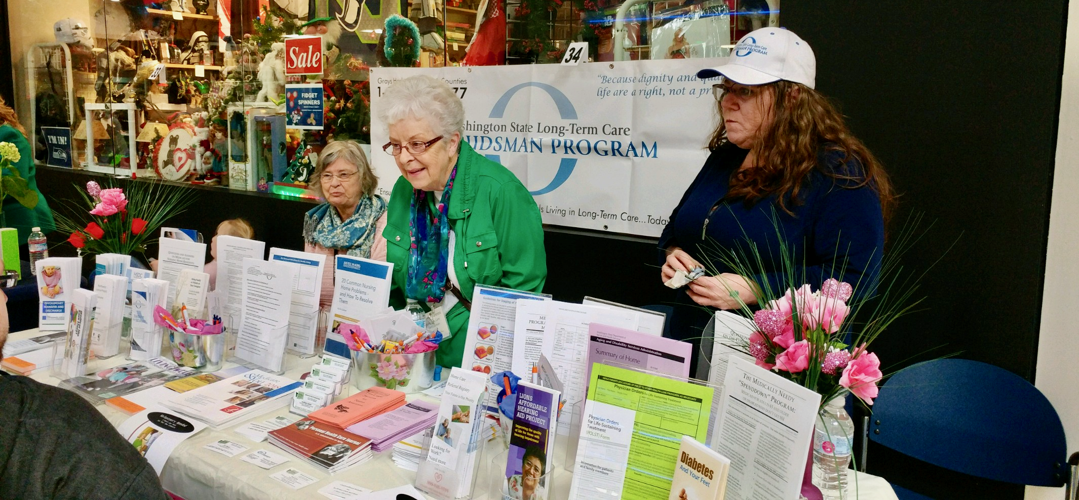 A group of three woman are standing behind an informational booth for the Washington State Long-Term Care Ombudsman Program. One woman is wearing a white hat with the logo, and the other two are speaking to people not pictured on the other side of the table.
