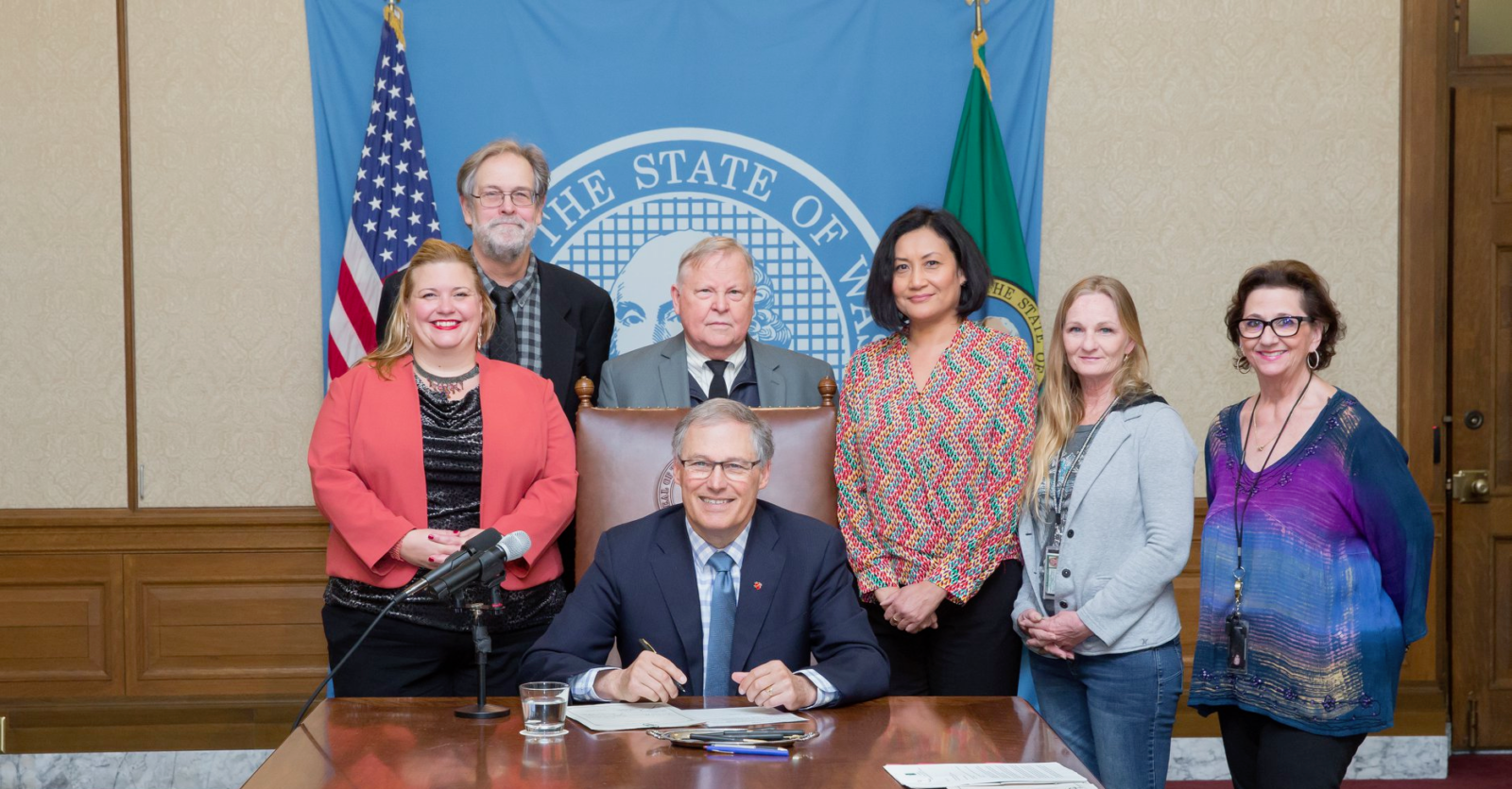 A group of people standing in a half semi-circle around the governor's desk. Behind the group is the blue State of Washington seal, a USA flag, and the Washington State flag. Governor Inslee is smiling at the camera and has a half-poised pen above a piece of legislation. The group is dressed semi-professionally. The group features Patricia Hunter the Washington State Long-Term Care Ombuds.